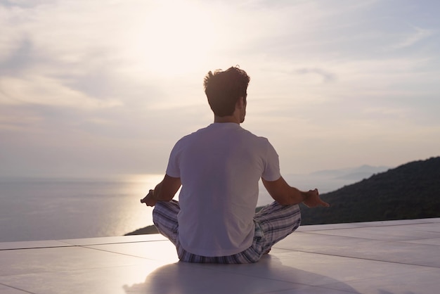 apuesto joven practicando yoga en la moderna terraza de la casa con el océano y la puesta de sol en el fondo