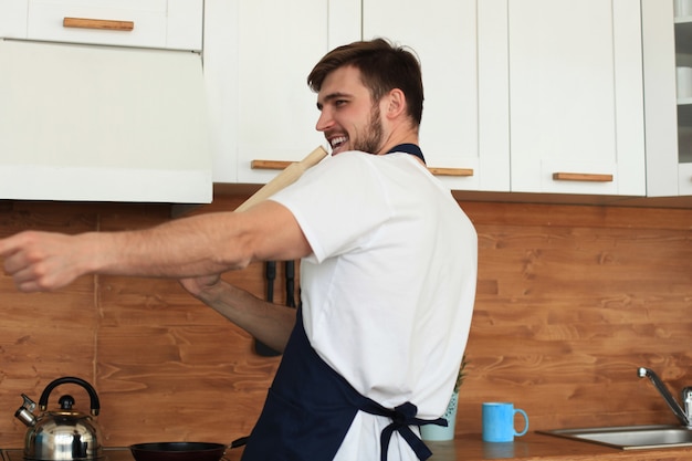 Apuesto joven en ropa casual cocinando el desayuno y sonriendo mientras está de pie en la cocina de su casa.