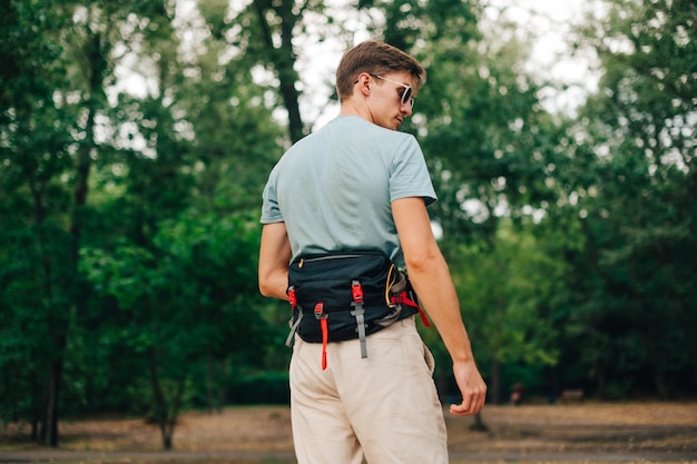 Apuesto joven en ropa casual de verano posando en el bosque con una bolsa de senderismo