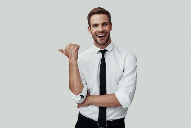 Apuesto joven en ropa formal apuntando copia espacio y sonriendo mientras está de pie contra el fondo gris