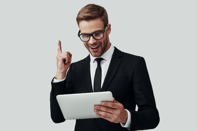 Apuesto joven en ropa formal trabajando con tableta digital y sonriendo mientras está de pie contra el fondo gris