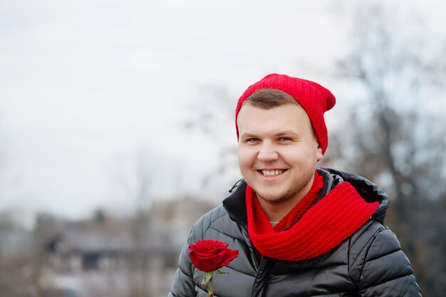 Apuesto joven con rosa roja en manos al aire libre en tiempo de nieve