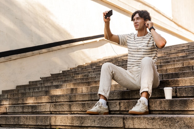 Foto apuesto joven sentado en pasos al aire libre escuchando música tomar un selfie por teléfono móvil.