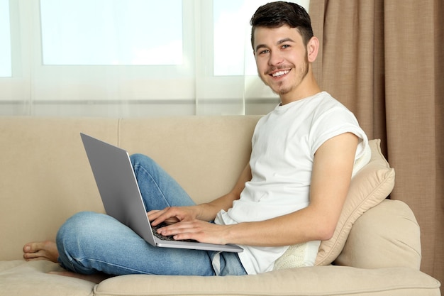 Apuesto joven sentado en el sofá y usando la computadora portátil en la habitación