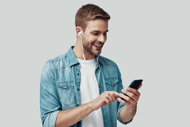 Apuesto joven sonriendo y escuchando música mientras está de pie contra el fondo gris