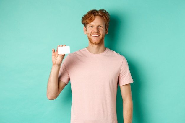 Apuesto joven sonriendo y mostrando tarjeta de crédito plástica, de pie en camiseta contra el fondo turquesa