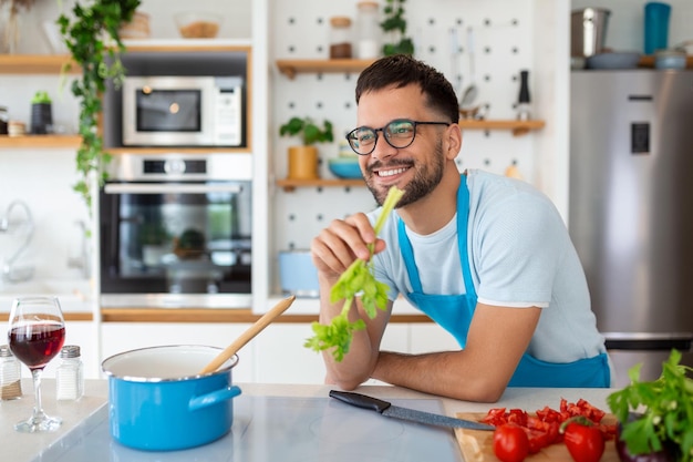 Apuesto joven sonriente apoyado en el mostrador de la cocina con verduras y mirando hacia otro lado