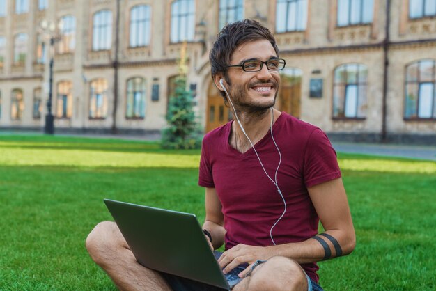 Apuesto joven sonriente usando computadora portátil mientras está sentado en el parque de la universidad