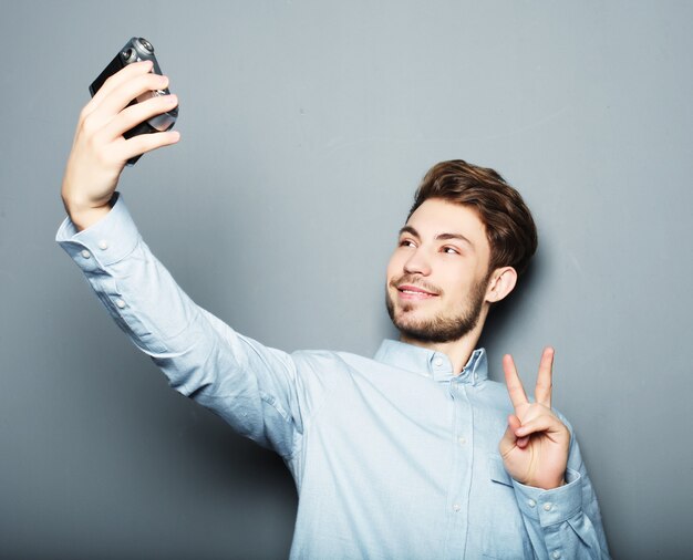 Apuesto joven sosteniendo la cámara y haciendo selfie y sonriendo mientras está de pie contra el espacio gris