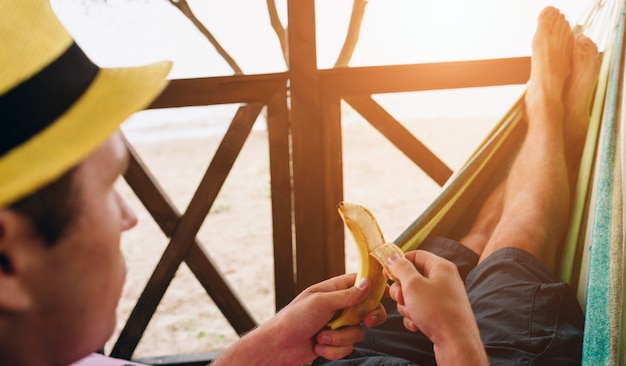 Apuesto joven sosteniendo plátano y acostado en una hamaca en una playa soleada junto a un océano