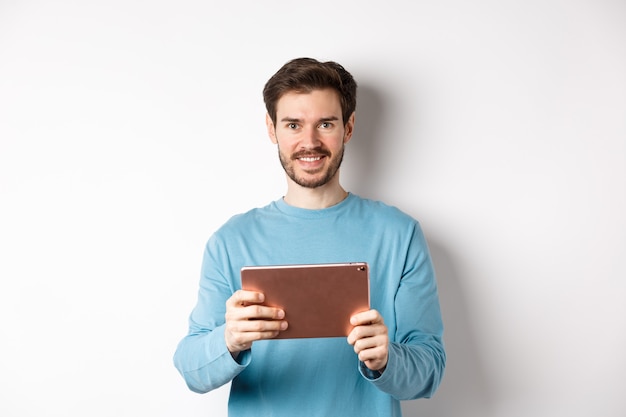 Apuesto joven sosteniendo tableta digital y sonriendo, redes con dispositivo, de pie sobre fondo blanco. Copia espacio