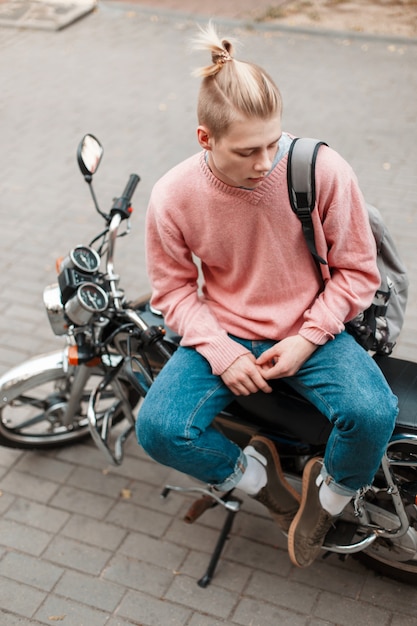 Apuesto joven con un suéter rosa y jeans con una mochila y con una patineta sentado en una motocicleta