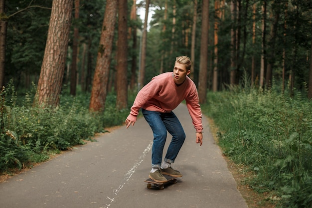 Apuesto joven con un suéter rosa y pantalones vaqueros azules monta una patineta sobre el asfalto en el parque