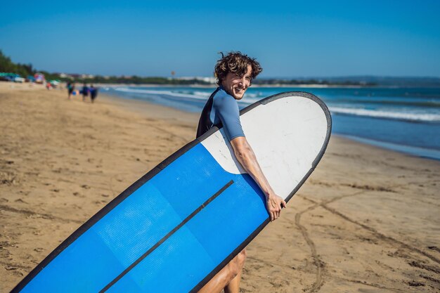 Apuesto joven surfista deportivo posando con su tabla de surf bajo el brazo en su traje de neopreno en una playa tropical de arena