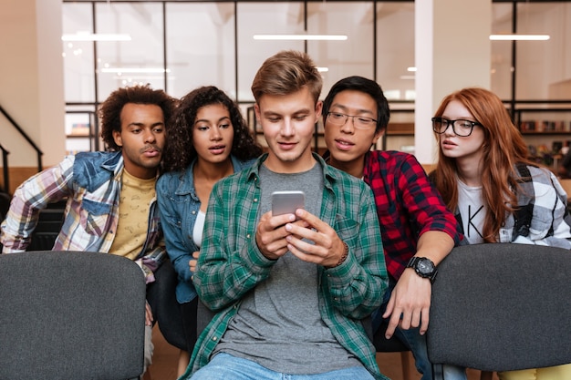 Apuesto joven y sus amigos sentados y usando el teléfono celular juntos