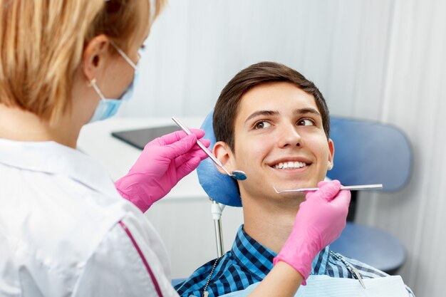 Apuesto joven tiene chequeo dental en consultorio dental. El dentista está examinando los dientes de un paciente con herramientas dentales. Odontología