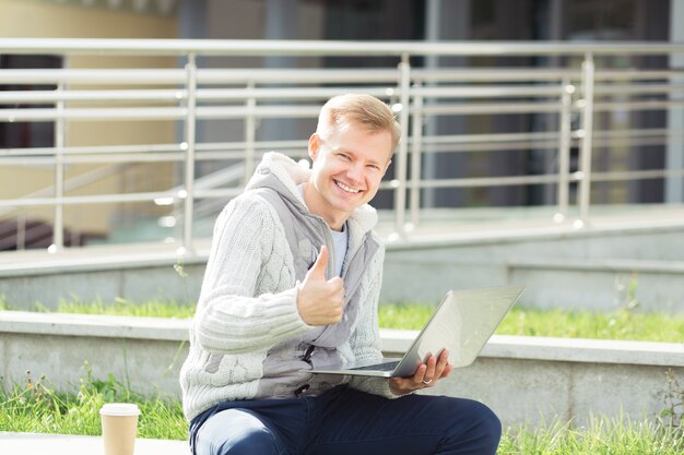 Apuesto joven trabajando en equipo portátil y sonriendo en la calle de la ciudad