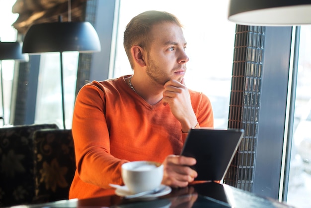 Apuesto joven trabajando con tableta pensando mirando por la ventana mientras disfruta de un café en la cafetería