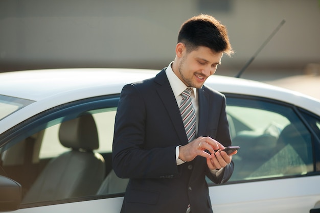 apuesto joven en traje cerca del coche con el teléfono en las manos