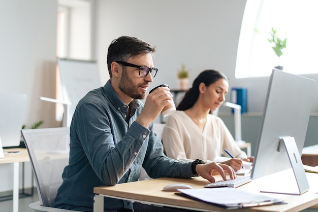 Foto apuesto joven usando computadora bebiendo café trabajando con su colega en espacio abierto