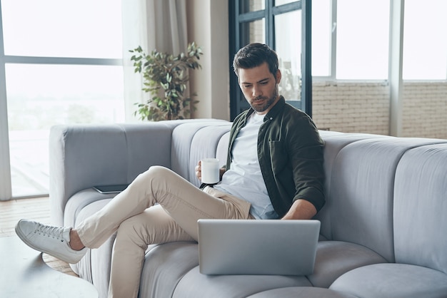 Apuesto joven usando una computadora portátil y tomando café mientras está sentado en el sofá en casa