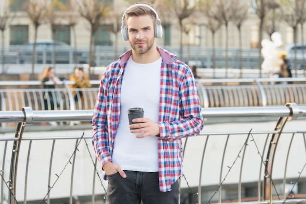 Apuesto joven usar auriculares casual vestido hombre bebiendo café de la mañana de la taza sin afeitar chico con cerdas escuchar música estilo urbano vida moderna concepto estudiante beber té