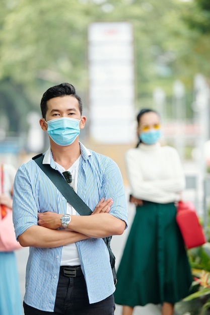 Apuesto joven vietnamita confiado en máscara médica de pie al aire libre con los brazos cruzados y mirando a la cámara