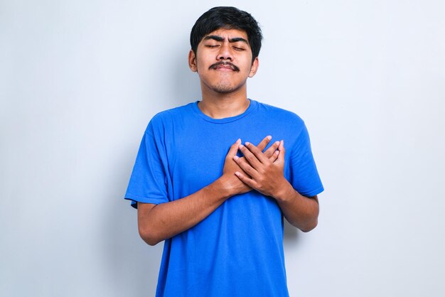 Apuesto joven vistiendo camiseta casual sonriendo con las manos en el pecho con los ojos cerrados y gesto de agradecimiento en la cara sobre fondo blanco. concepto de salud.