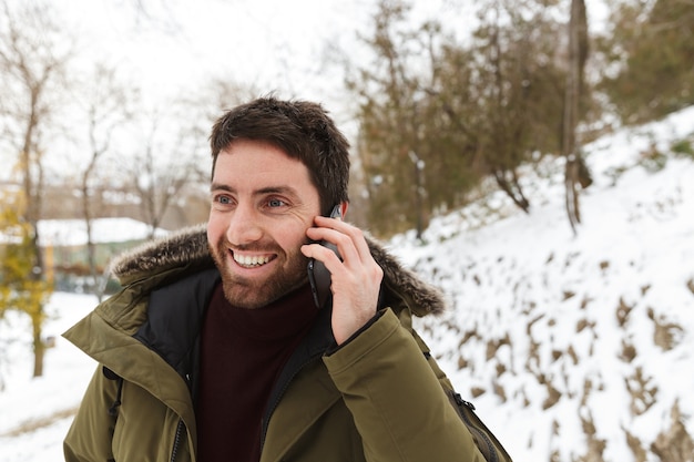 Apuesto joven vistiendo chaqueta de invierno de pie al aire libre, hablando por teléfono móvil