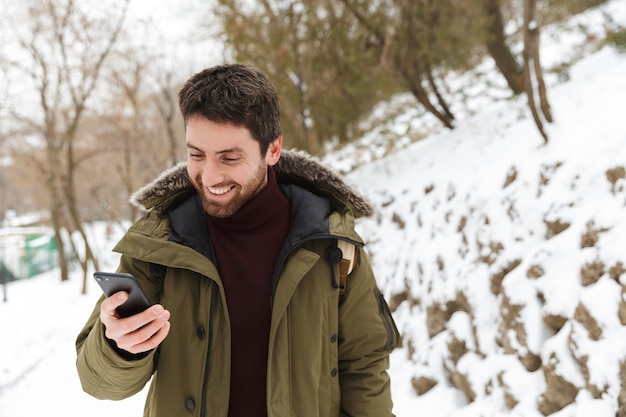 Apuesto joven vistiendo chaqueta de invierno mediante teléfono móvil mientras camina al aire libre