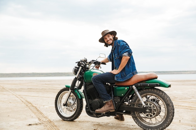 Apuesto joven vistiendo ropa casual sentado en una motocicleta en la playa.