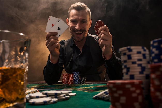 Apuesto jugador de póquer con dos ases en sus manos y fichas sentado en la mesa de póquer en una habitación oscura llena de humo de cigarrillo.