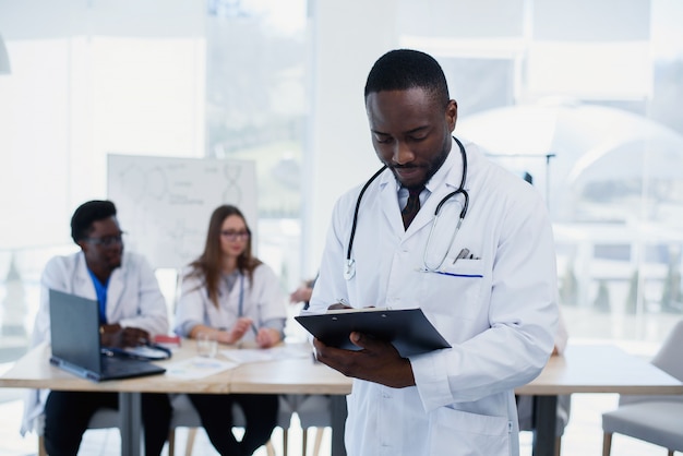 Apuesto médico africano en bata blanca hace algunas notas. Joven estudiante de medicina con un estetoscopio alrededor del cuello tiene una carpeta.