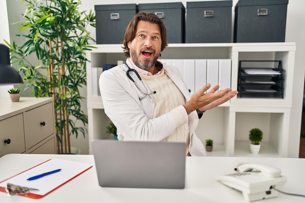 Apuesto médico de mediana edad que trabaja en la clínica señalando a un lado con las manos abiertas mostrando el espacio de copia presentando un anuncio sonriendo emocionado feliz