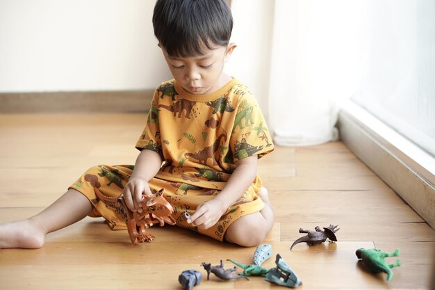 Apuesto niño asiático felizmente jugando con un juguete de dinosaurio de plástico cerca de la ventana en un hotel