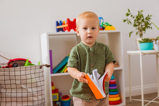 El apuesto niño pequeño está parado en medio de una sala de juegos con un libro