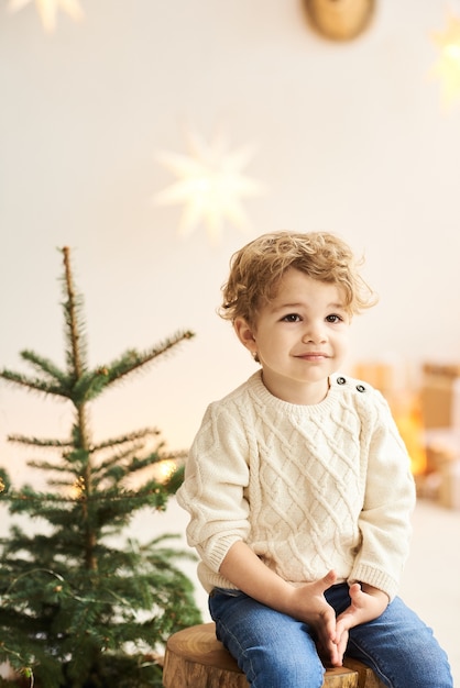 Un apuesto niño rizado se sienta en una silla de madera cerca de un árbol de Navidad en una habitación blanca