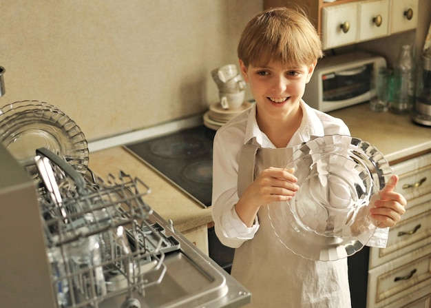 Un apuesto niño sonriente de 10 años hace las tareas del hogar descargando platos limpios del lavavajillas