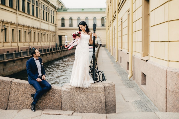 Apuesto novio se sienta en el puente mira a su bella novia que se pone de pie y le dice cumplidos