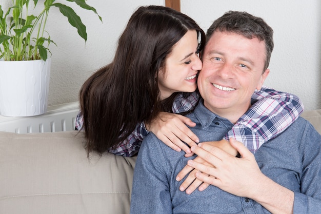 Apuesto padre alegre sentado en el sofá de casa sonriendo y su hija dulce beso