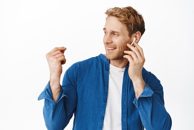 Un apuesto pelirrojo escucha música con auriculares inalámbricos mirando a un lado el logo de la pancarta sonriendo complacido tocando el auricular en la oreja y bailando de pie sobre fondo blanco