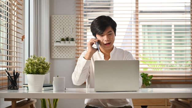Apuesto trabajador independiente masculino asiático mirando la pantalla del portátil y hablando por teléfono móvil