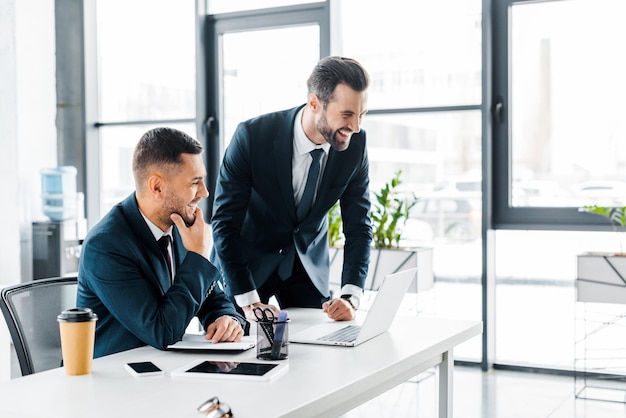 Apuestos hombres de negocios riéndose cerca de la computadora portátil en la oficina moderna