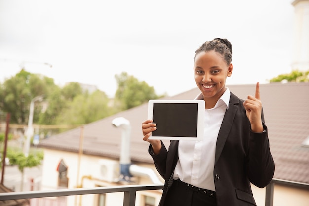 Apuntando hacia arriba con la tableta. Empresaria afroamericana en traje de oficina sonriendo, parece segura y feliz. Concepto de finanzas, negocios, igualdad y derechos humanos. Hermosa joven modelo, exitosa.