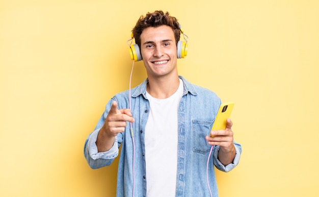 Apuntando a la cámara con una sonrisa satisfecha, confiada y amistosa, eligiéndote. concepto de auriculares y teléfono inteligente