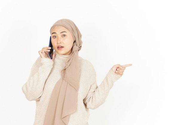 Apuntando a un lado y hablando por teléfono de la hermosa mujer asiática vistiendo Hijab aislado en blanco