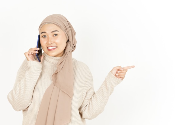 Apuntando a un lado y hablando por teléfono de la hermosa mujer asiática vistiendo Hijab aislado en blanco