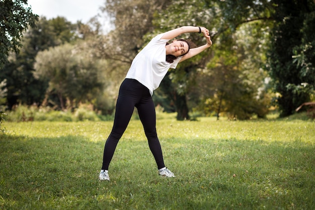 Foto aquecimento no parque. uma jovem apta mulher caucasiana em roupas esportivas, fazendo flexões laterais. o conceito de treinamento esportivo