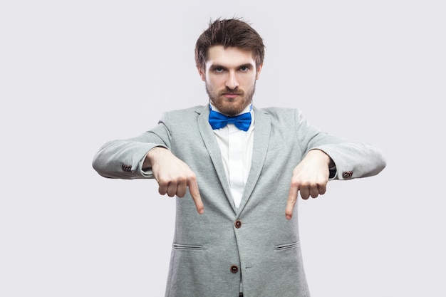 Aquí y ahora mismo. Retrato de hombre barbudo guapo mandón serio en traje gris casual y pajarita azul de pie apuntando y mirando a cámara. tiro del estudio de interior, aislado en fondo gris claro.