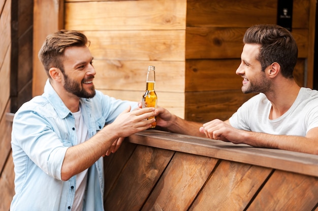 ¡Aquí tienes tu cerveza! Camarero joven feliz dando una botella de cerveza a su cliente mientras está de pie en la barra del bar